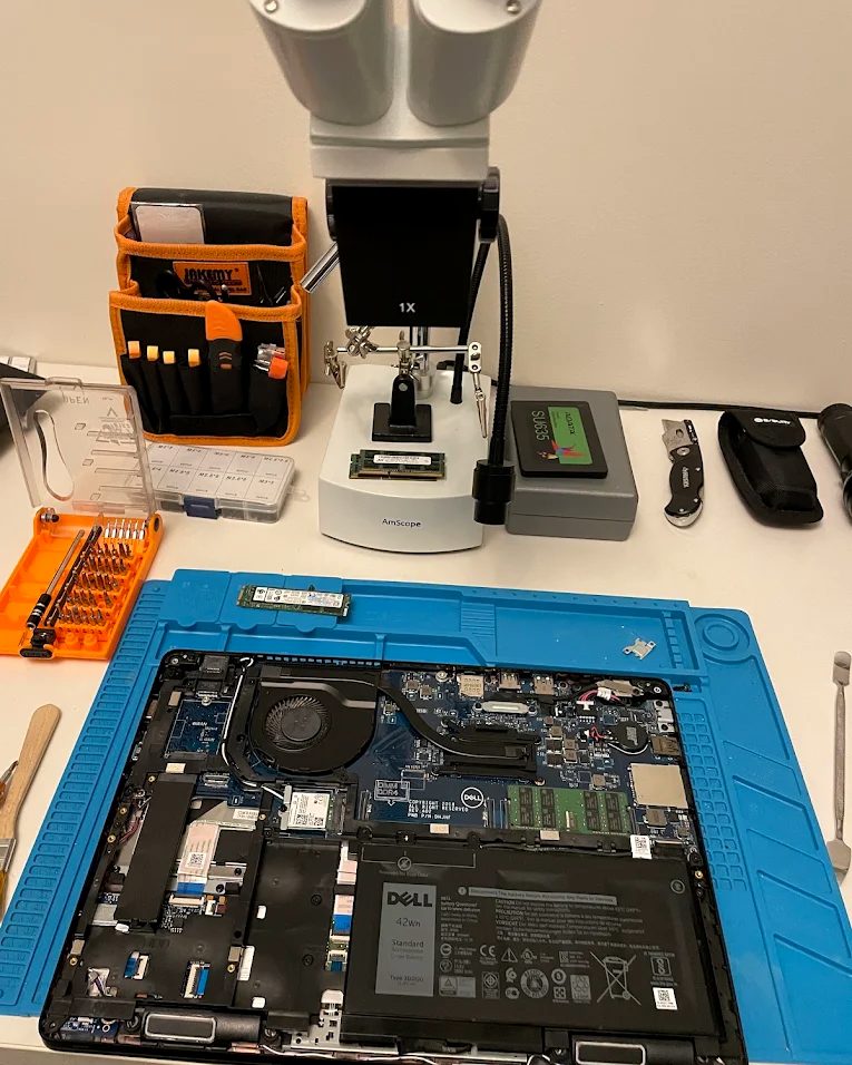 A Dell laptop opened on an anti-static mat for repair, with a microscope, replacement SSD and precision tools on the bench